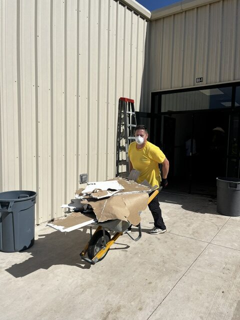 A volunteer wearing a yellow shirt and a mask hauls a wheelbarrow full of debris out from a building.