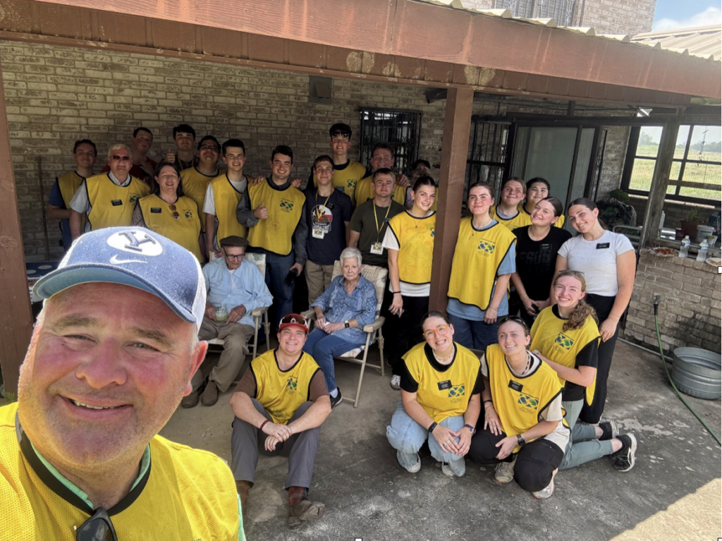 The image shows a group of volunteers gathered in an outdoor setting, with some wearing yellow shirts and others in different attire, under a brick wall structure. Home owners are seen with them.